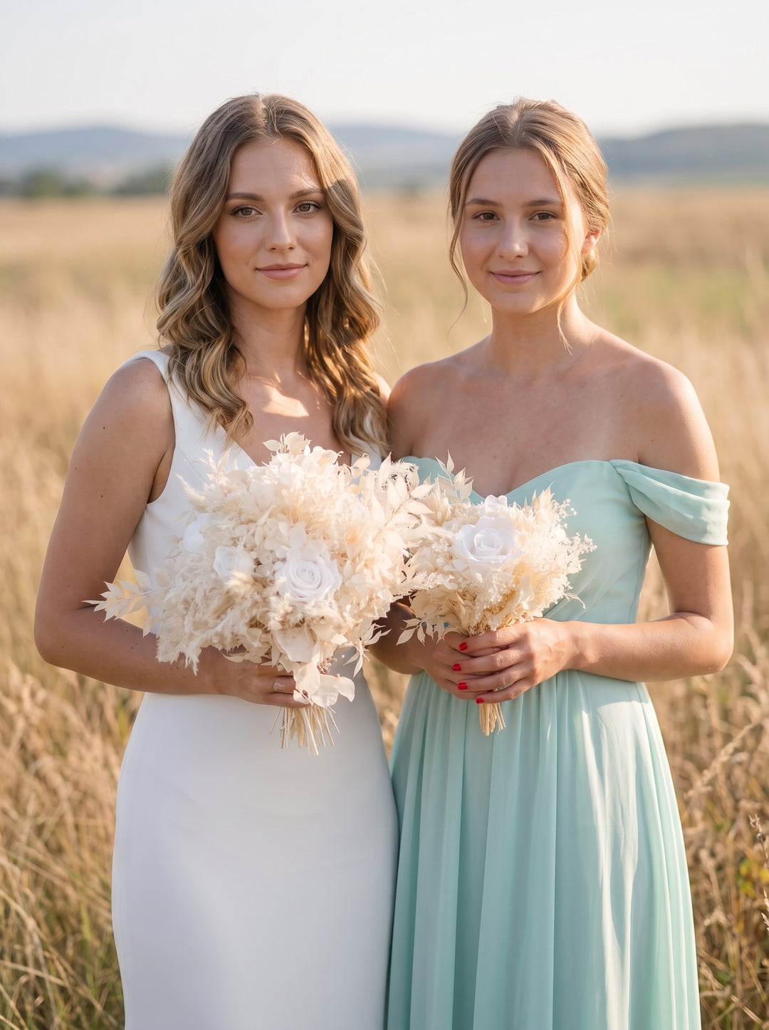 Two women standing in a sunlit field, each holding a large cream floral bouquet. The woman on the left wears a sleeveless white dress, and the woman on the right wears an off-the-shoulder mint green dress.