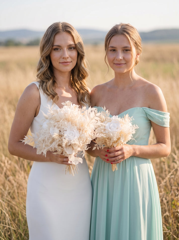 Two women standing in a sunlit field, each holding a large cream floral bouquet. The woman on the left wears a sleeveless white dress, and the woman on the right wears an off-the-shoulder mint green dress.