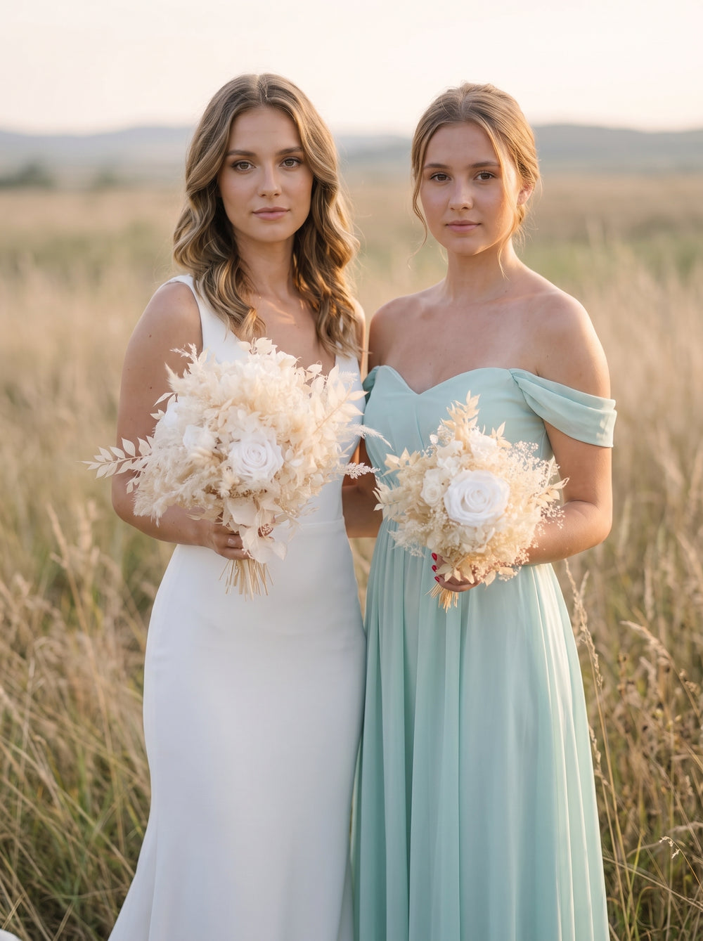 Two women standing in a sunlit field, each holding a large dried floral bouquet. One wears a sleeveless white wedding gown, and the other wears an off-the-shoulder mint green bridesmaid dress.