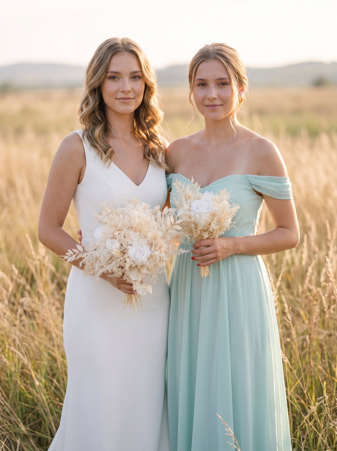 Two women standing in a sunlit field of tall dry grass, wearing floor-length bridesmaid-style dresses. One dress is white and sleeveless with a V-neckline, and the other is a soft off-the-shoulder mint or pale aqua dress.