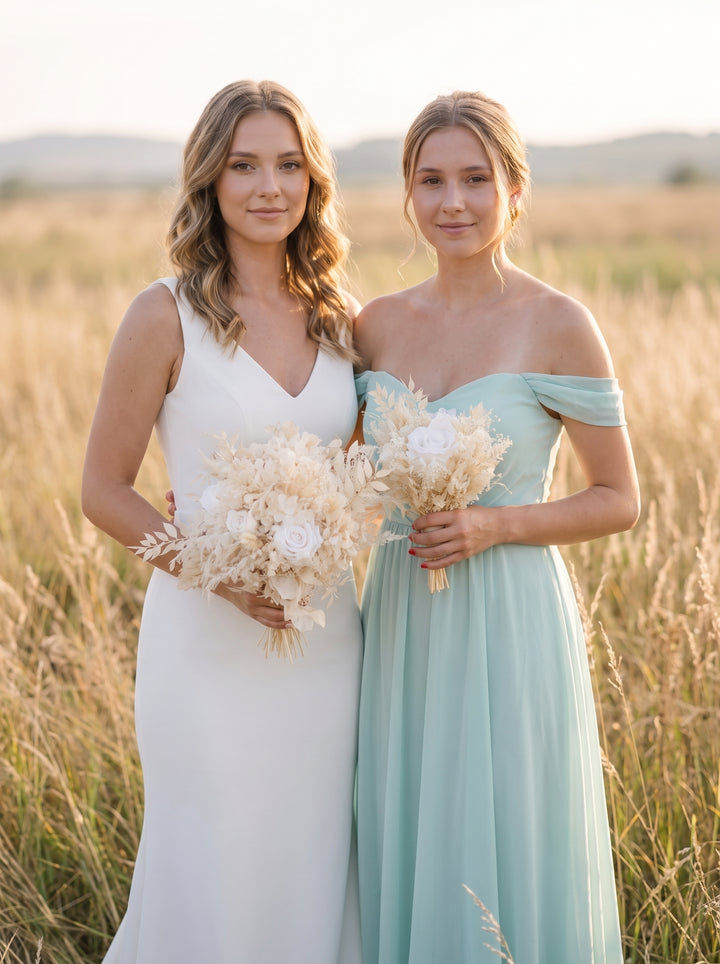 Two women standing in a sunlit field of tall dry grass, wearing floor-length bridesmaid-style dresses. One dress is white and sleeveless with a V-neckline, and the other is a soft off-the-shoulder mint or pale aqua dress.