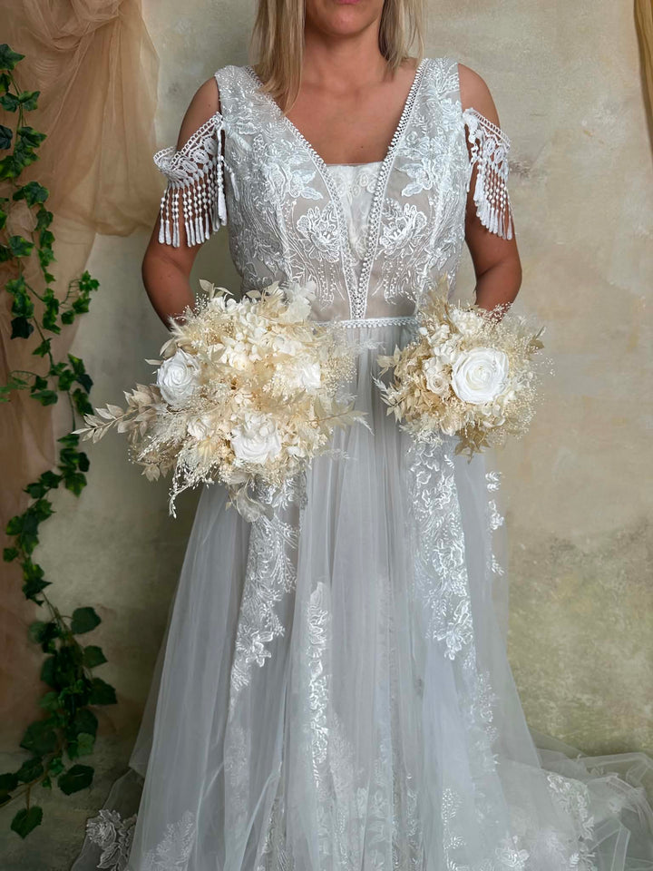 A woman wearing a white lace cold-shoulder wedding gown with tulle skirt, holding two large cream floral bouquets against a neutral textured backdrop.