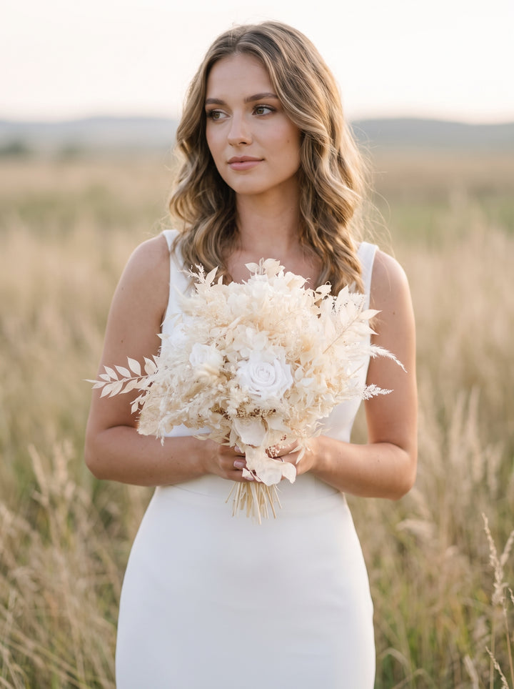 A bride standing in a sunlit field wearing a sleeveless white dress and holding a large bouquet of cream and white dried-style flowers.