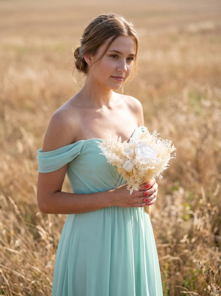 A bridesmaid standing in a dry grass field wearing an off-the-shoulder mint green dress and holding a bouquet of dried white flowers.