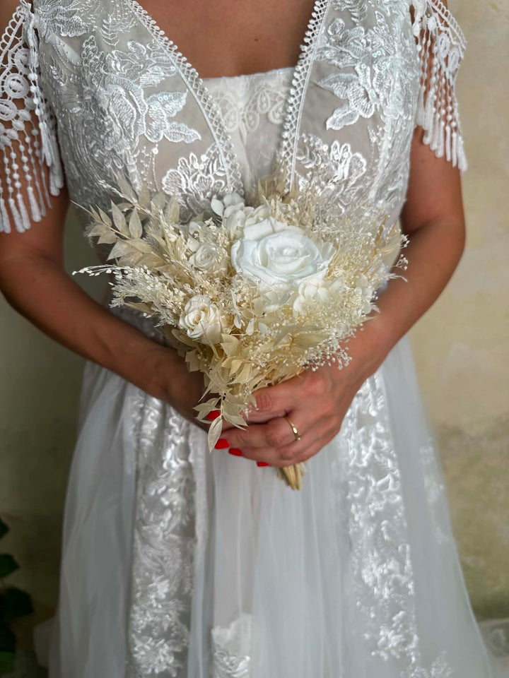 A close-up of a bride holding a dried floral bouquet in a white lace wedding dress with tassel fringe on the sleeves.