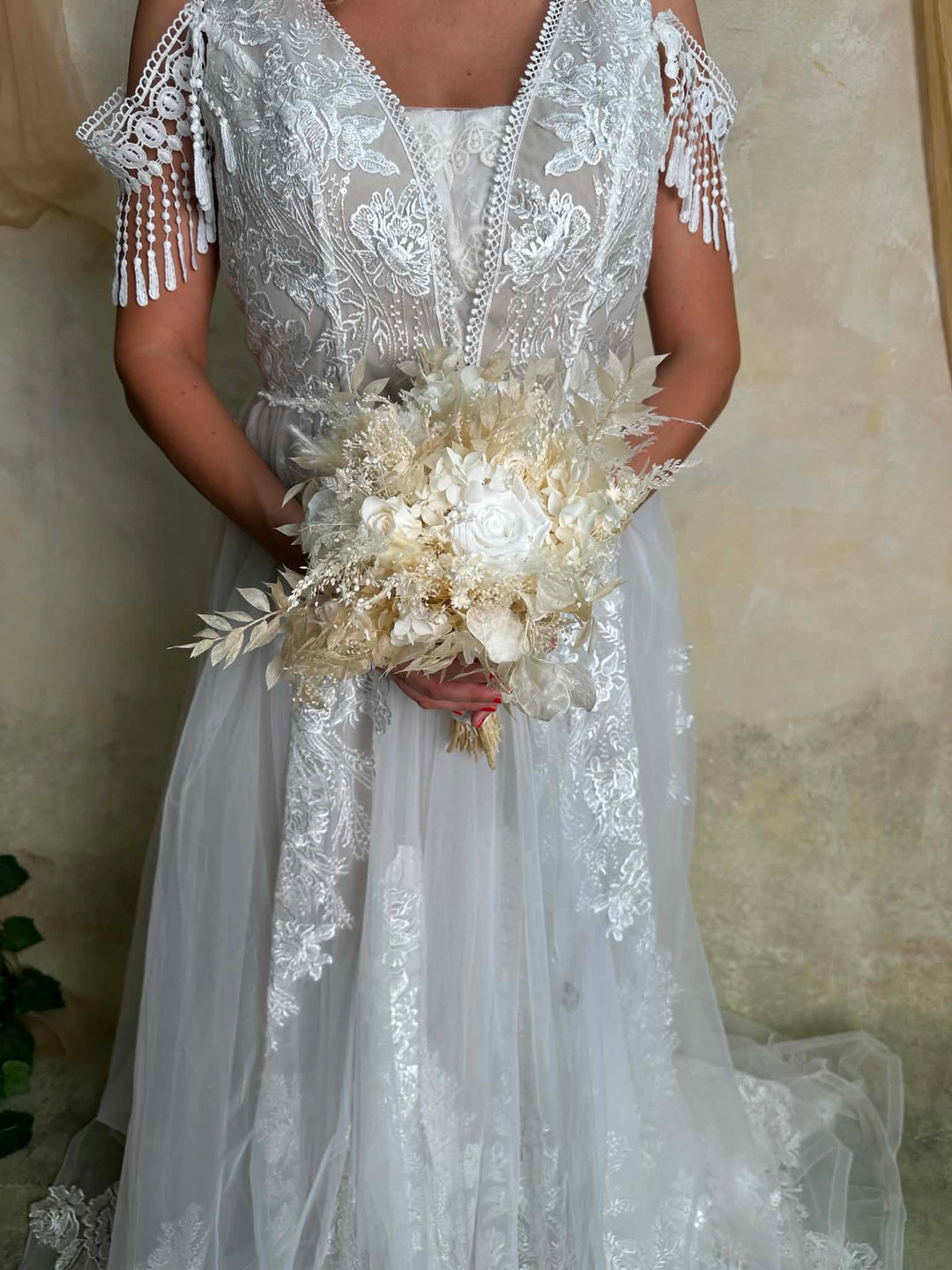 A woman wearing a white lace wedding dress with off-the-shoulder fringed sleeves and holding a bouquet of cream dried flowers, standing against a textured beige wall.