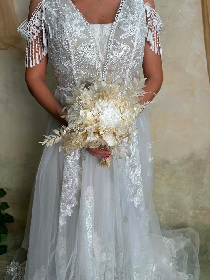 A woman wearing a white lace wedding dress with off-the-shoulder fringed sleeves and holding a bouquet of cream dried flowers, standing against a textured beige wall.