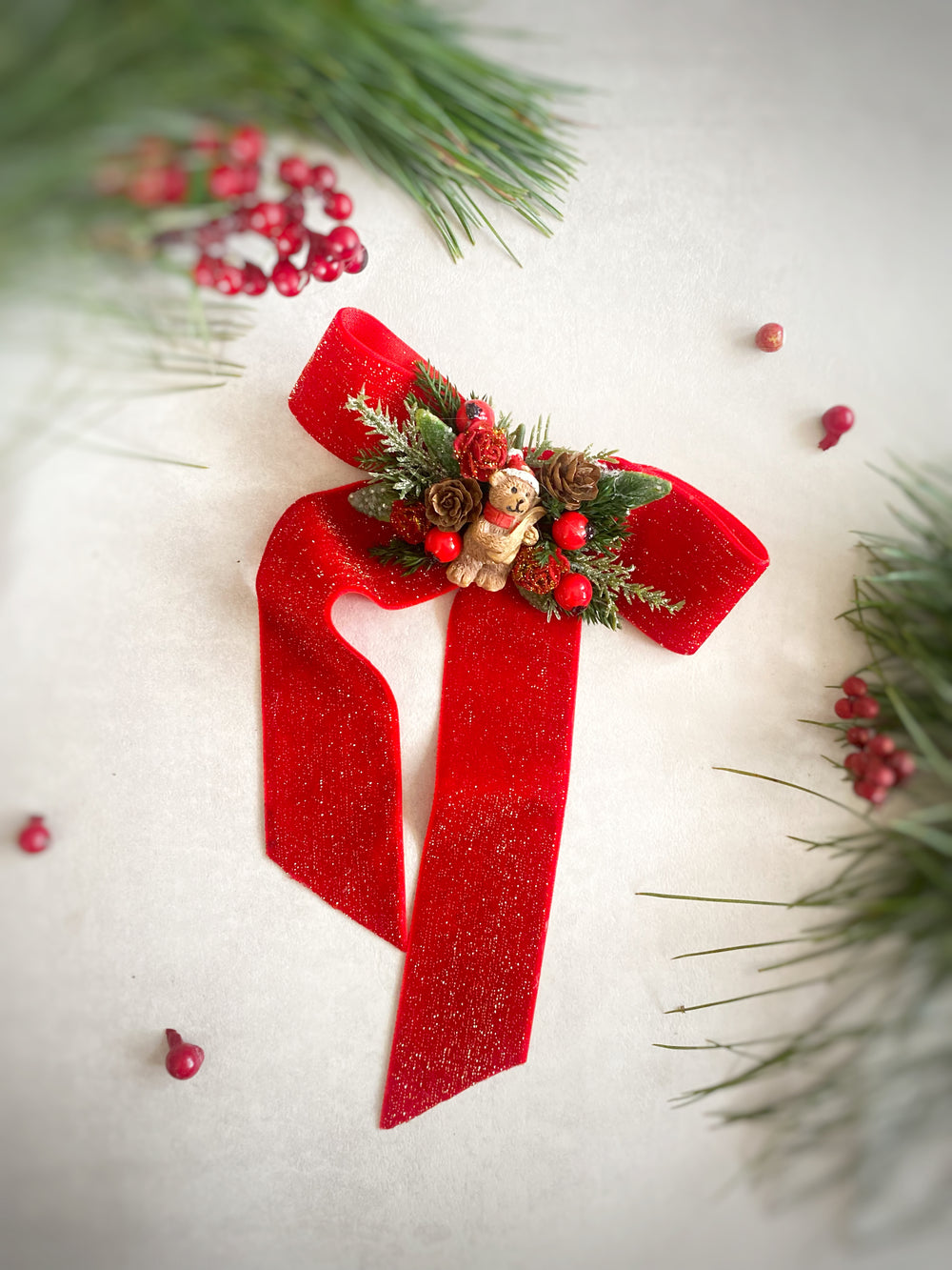 A festive red glitter ribbon bow decorated with a small teddy ornament, pinecones, and evergreen branches, arranged on a light cream background with holiday berries and pine needles.