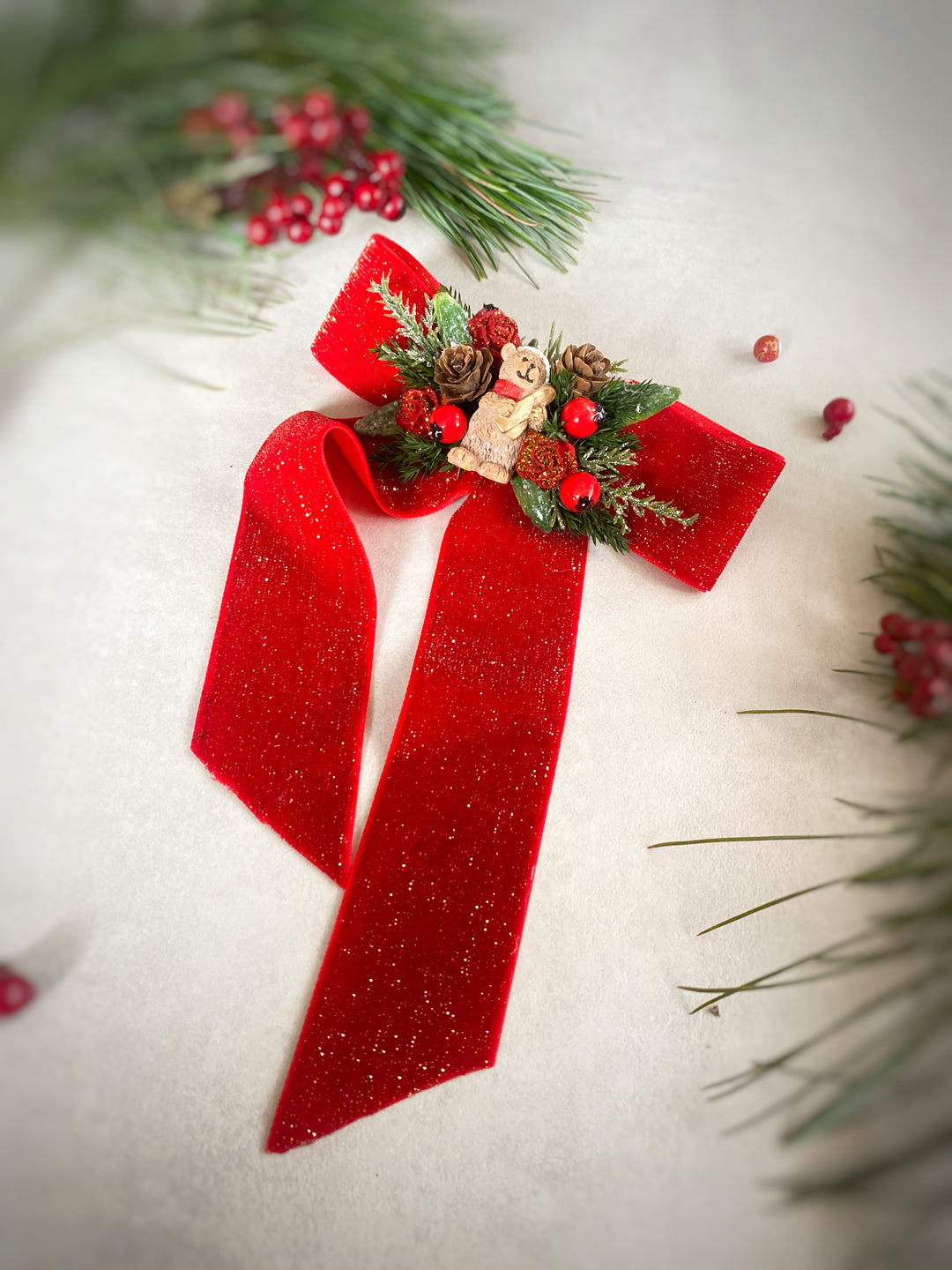 A festive Christmas bow decoration made of glittery red ribbon, decorated with green pine branches, red berries, pinecones, and a small teddy bear figure, arranged on a light textured background with holiday greenery.