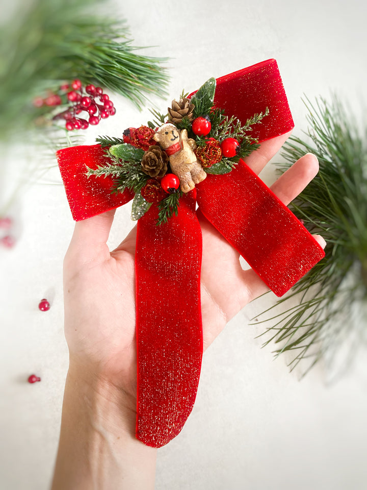 A hand holds a festive Christmas stocking ornament shaped like a red velvet ribbon stocking, decorated with green pine branches, red berries, pinecones, and a small teddy bear figure, against a soft white background with blurred pine needles.