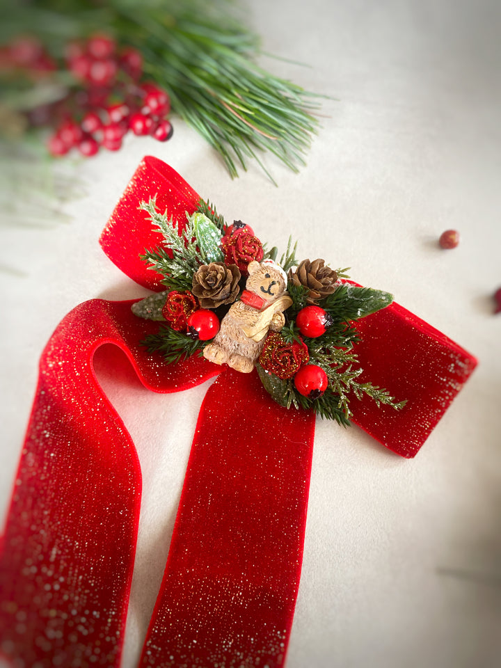 A festive Christmas decoration featuring a glittery red ribbon bow adorned with pinecones, red berries, green pine needles, and a small gingerbread man ornament, set against a light neutral background with blurred holiday greenery.