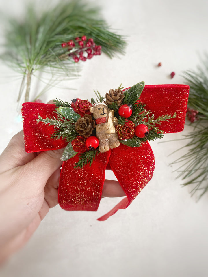 A hand holds a festive Christmas ornament shaped like a red glitter ribbon bow decorated with green pine branches, red berries, pinecones, and a small teddy bear wearing a red scarf, against a light background with blurred pine branches.