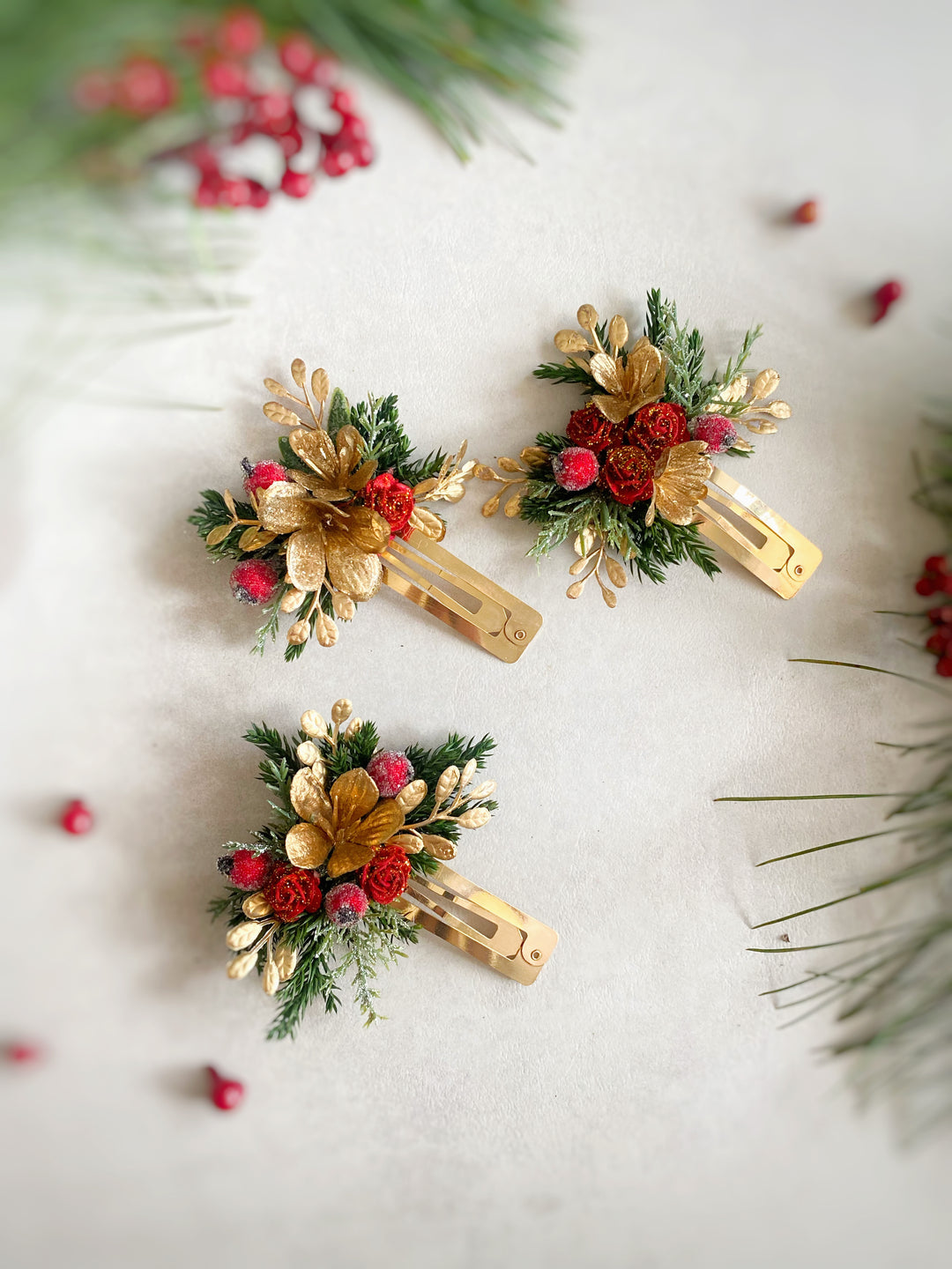 Three gold hair clips decorated with green pine needles, gold leaves, red berries, and small red roses, arranged on a light textured background with festive blurred pine branches.