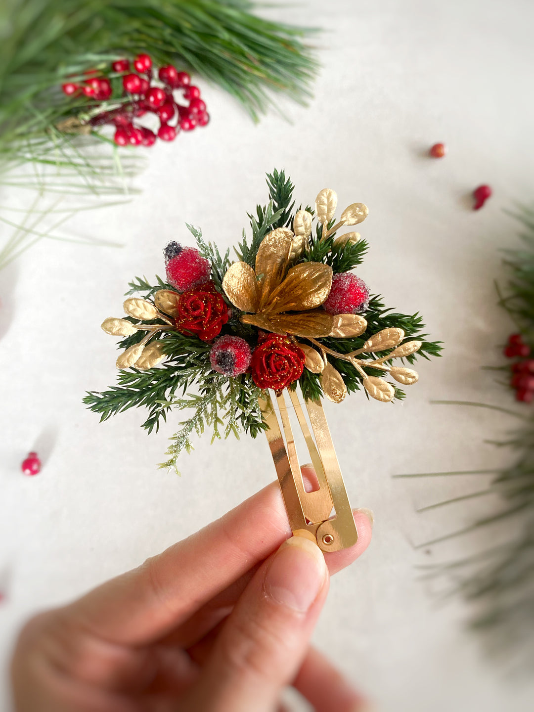 A hand holds a small decorative hair claw clip decorated with red berries, green pine needles, and gold metallic accents, set against a light background with blurred holiday greenery.