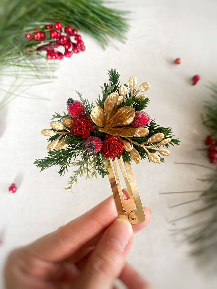 A hand holds a small decorative hair claw clip decorated with red berries, green pine needles, and gold metallic accents, set against a light background with blurred holiday greenery.