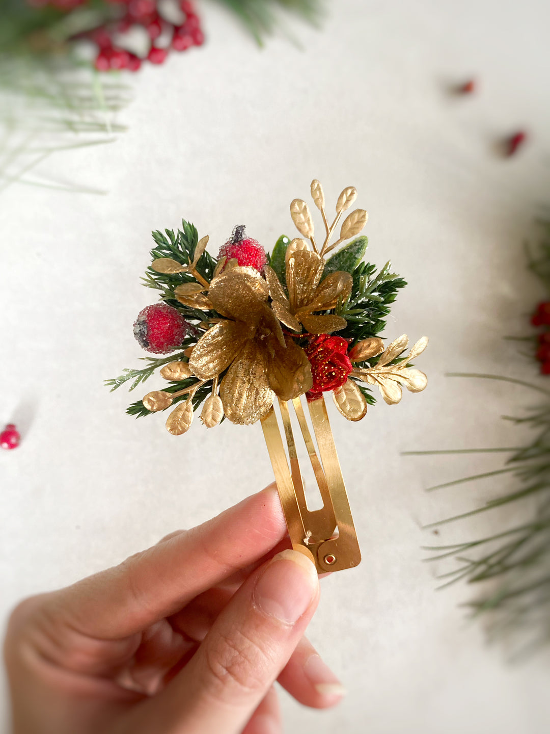 A hand holds a gold-tone hair claw clip decorated with a gold flower, green pine needles, and red berries, set against a soft, light background with blurred holiday greenery.