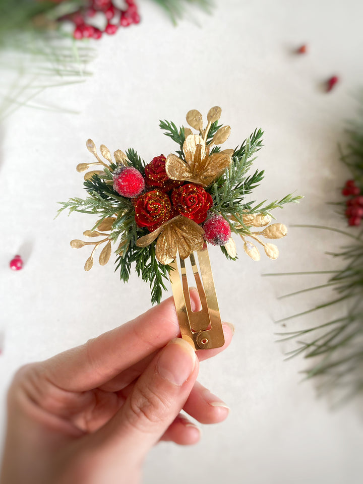 A hand holds a small gold hair claw clip decorated with green pine needles, red glittery berries, and gold leaf accents, against a softly blurred white background with holiday greenery.