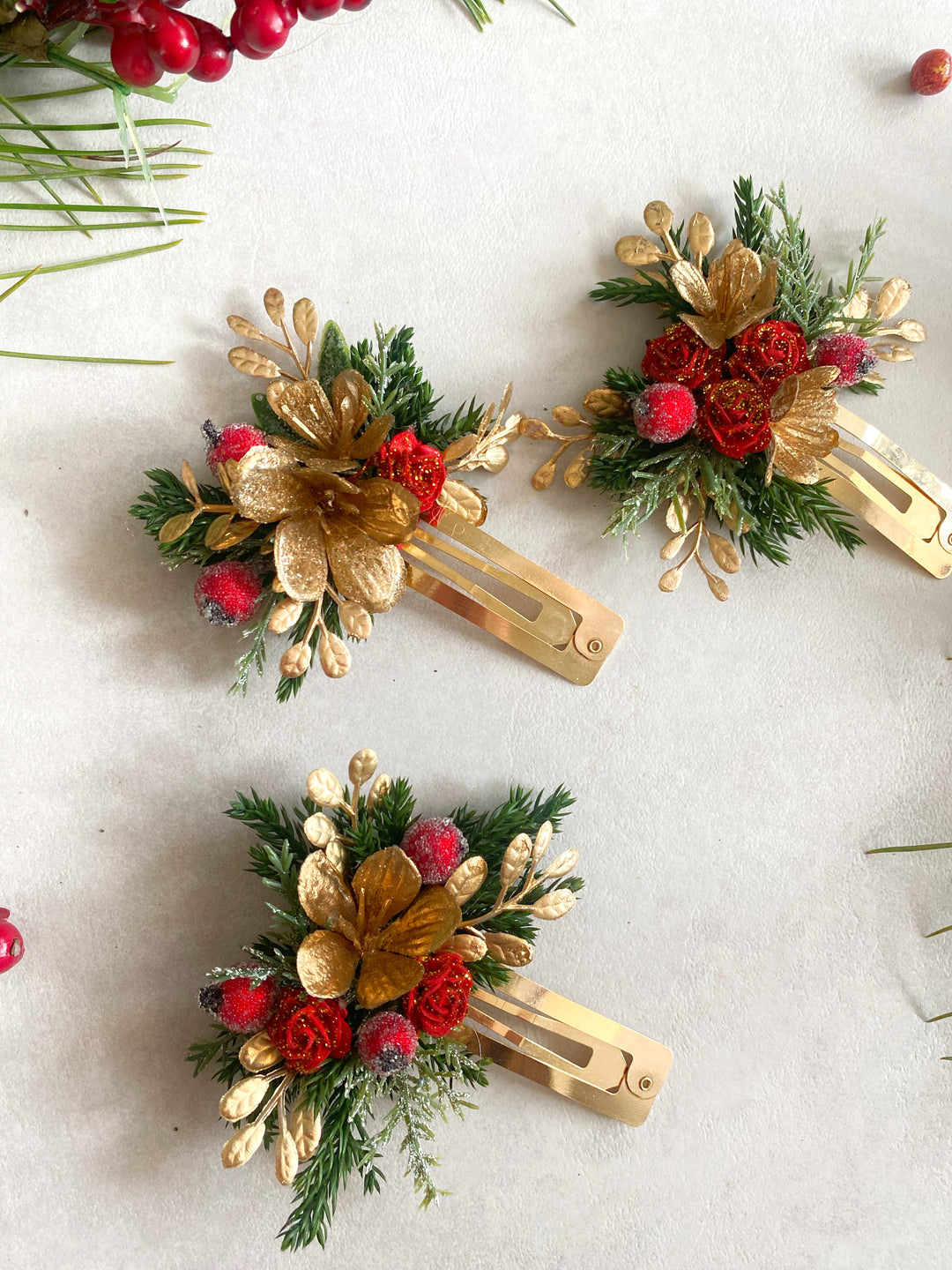 Three gold hair clips decorated with green pine branches, gold artificial leaves, red berries, and small white pinecones, arranged on a light beige background with holiday greenery.