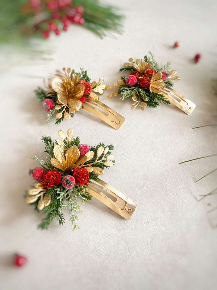 Three small gold hair clips decorated with needles, red berries, and green foliage, arranged on a light neutral surface.