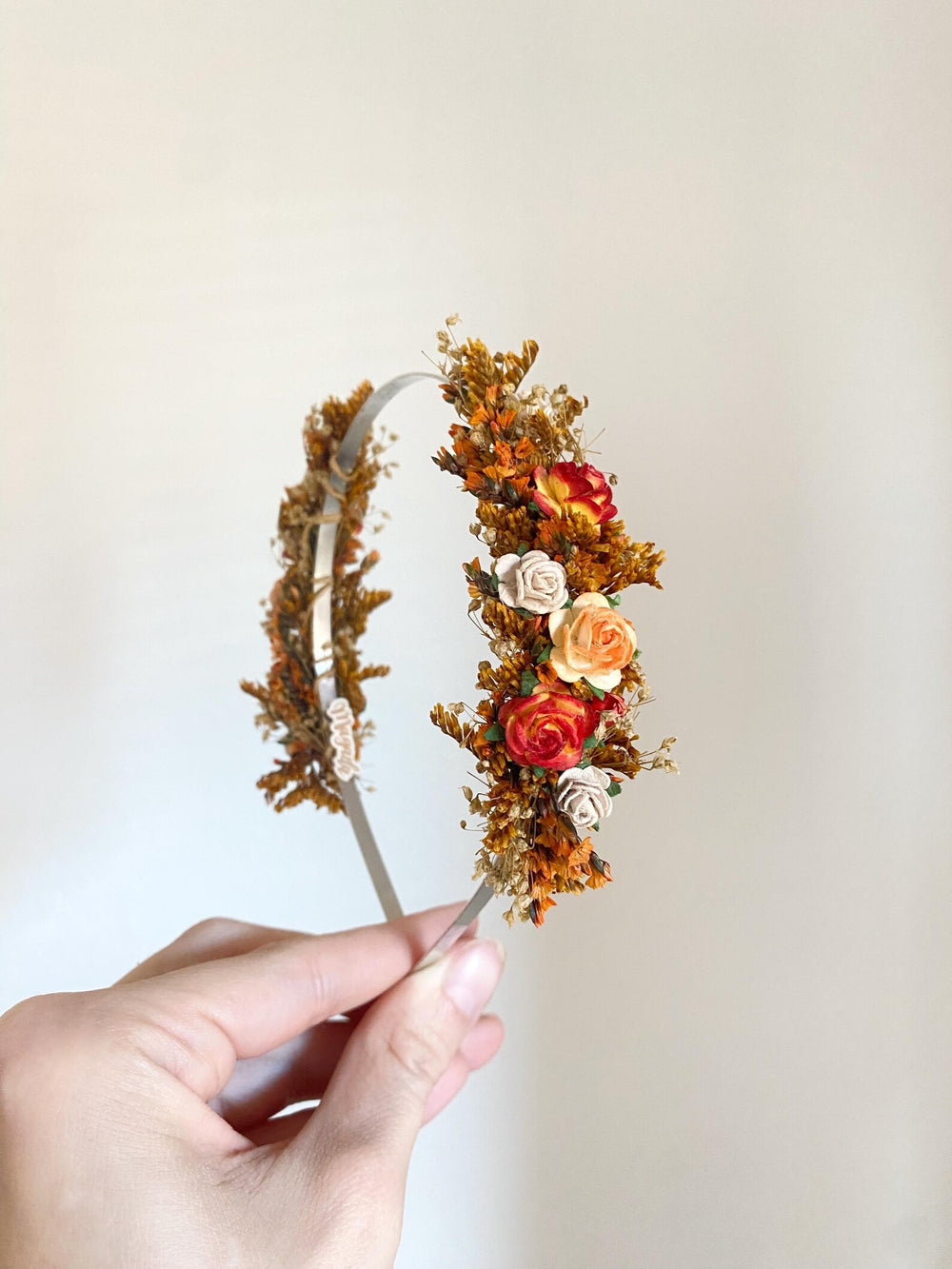 A hand holding a slim metal headband decorated with orange and brown botanicals and small artificial flowers in red, peach, and cream, against a plain light beige background.