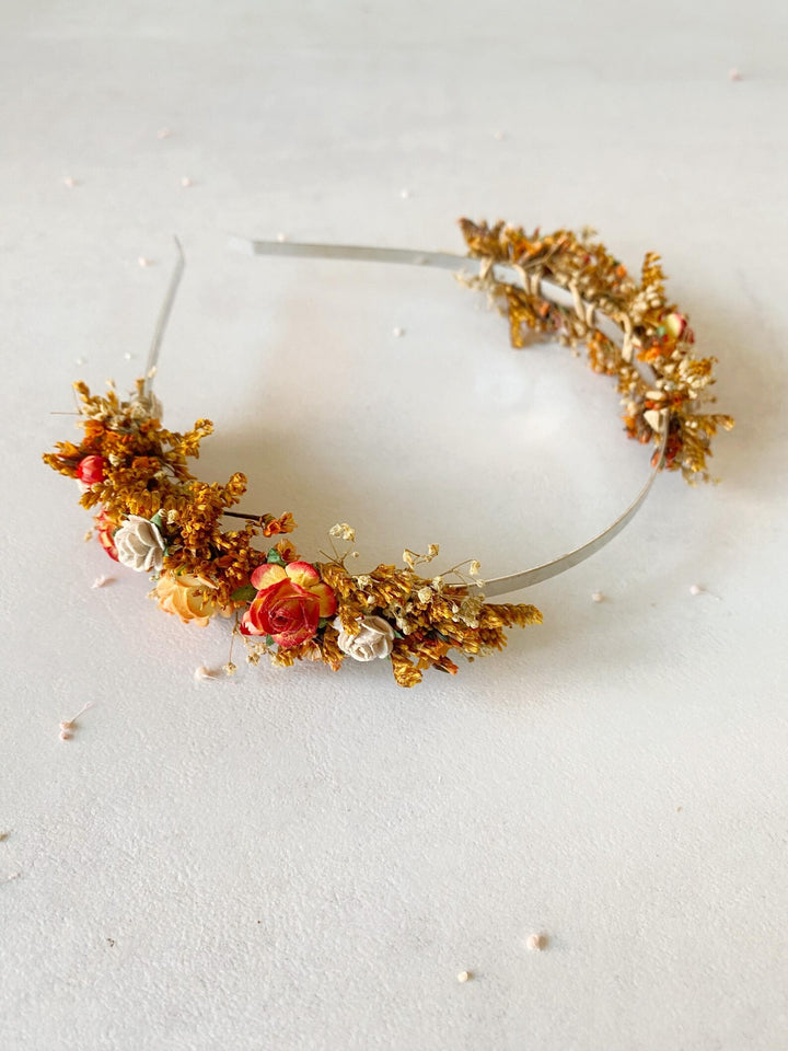 A delicate floral headband featuring orange and red flowers, small white blossoms, and textured gold leaf accents on a thin metallic band, displayed on a light off-white background.