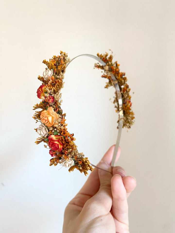 A hand holding a clear plastic headband decorated with orange and red flowers, yellow botanicals, and small white dried flowers against a plain off-white background.