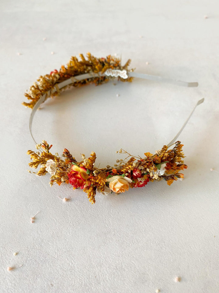 A floral headband featuring autumn flowers in shades of orange, red, yellow, and brown, attached to a thin clear elastic band, displayed on a light textured background.