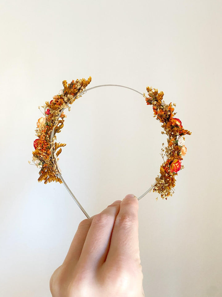 A floral crown headband with orange, brown, and red botanical elements on a thin metal band, held by a hand against a plain off-white background.