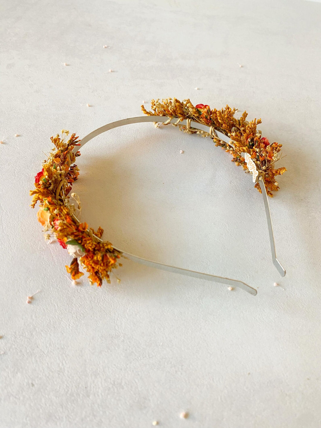 A silver metal headband decorated with orange and yellow flowers, small red blossoms, and white pearl-like accents, photographed on a light off-white textured background.