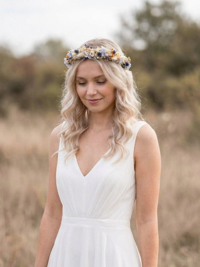 A woman wearing a sleeveless white V-neck maxi dress and a floral crown stands in a dry grassy field outdoors.