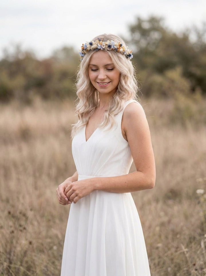 A woman wearing a sleeveless white V-neck dress and a floral crown stands outdoors in a dry grassy field, looking down with a gentle smile.