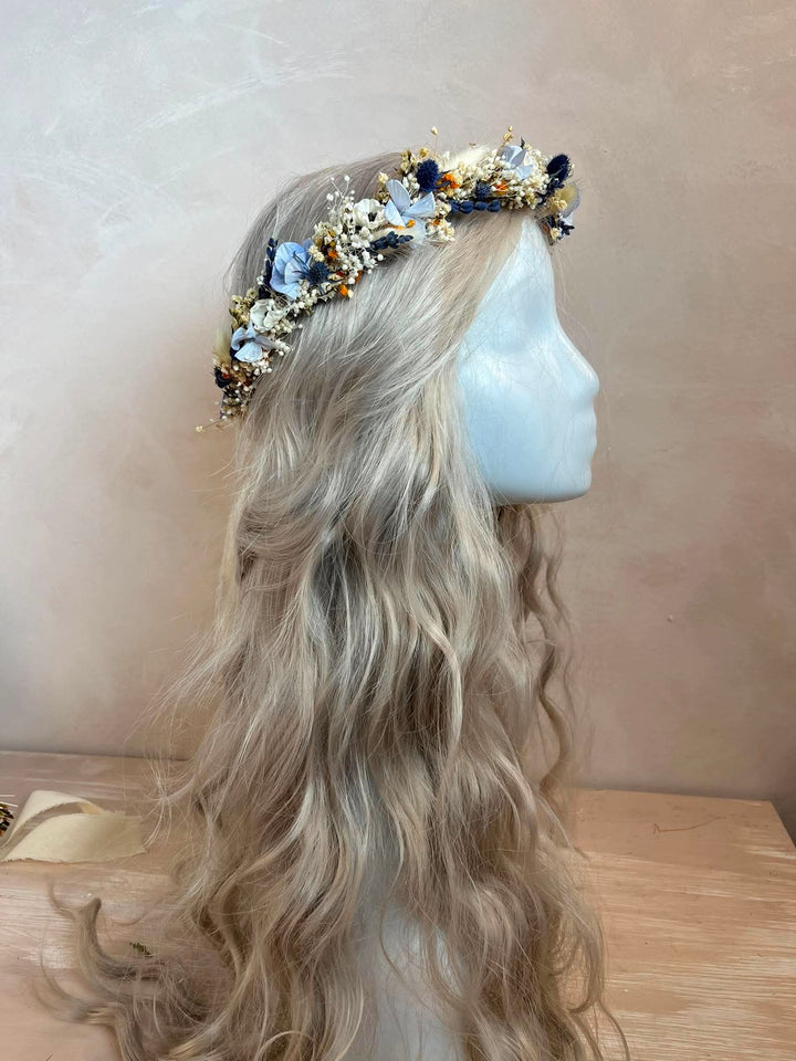A long wavy blonde wig displayed on a white mannequin head, styled with a floral crown featuring blue, gold, and white flowers against a neutral beige background.