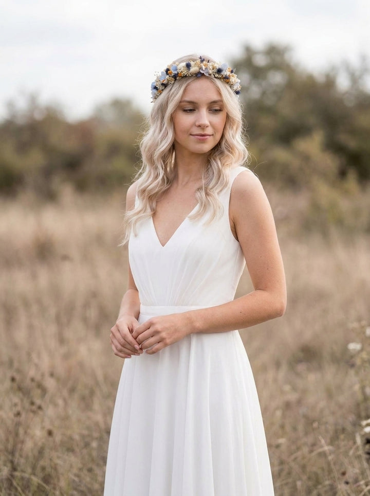 A woman standing in a dry grass field wearing a sleeveless white V-neck dress and a floral crown with blue and gold details.