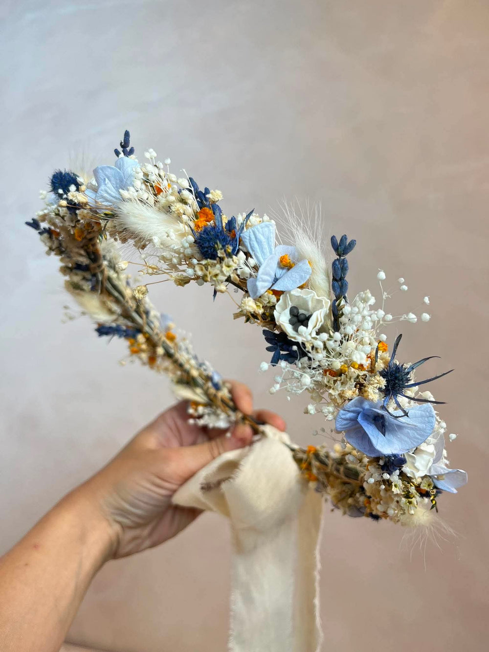 A dried floral headband with blue flowers, white baby's breath, and beige grass elements, held by a hand against a neutral beige background.
