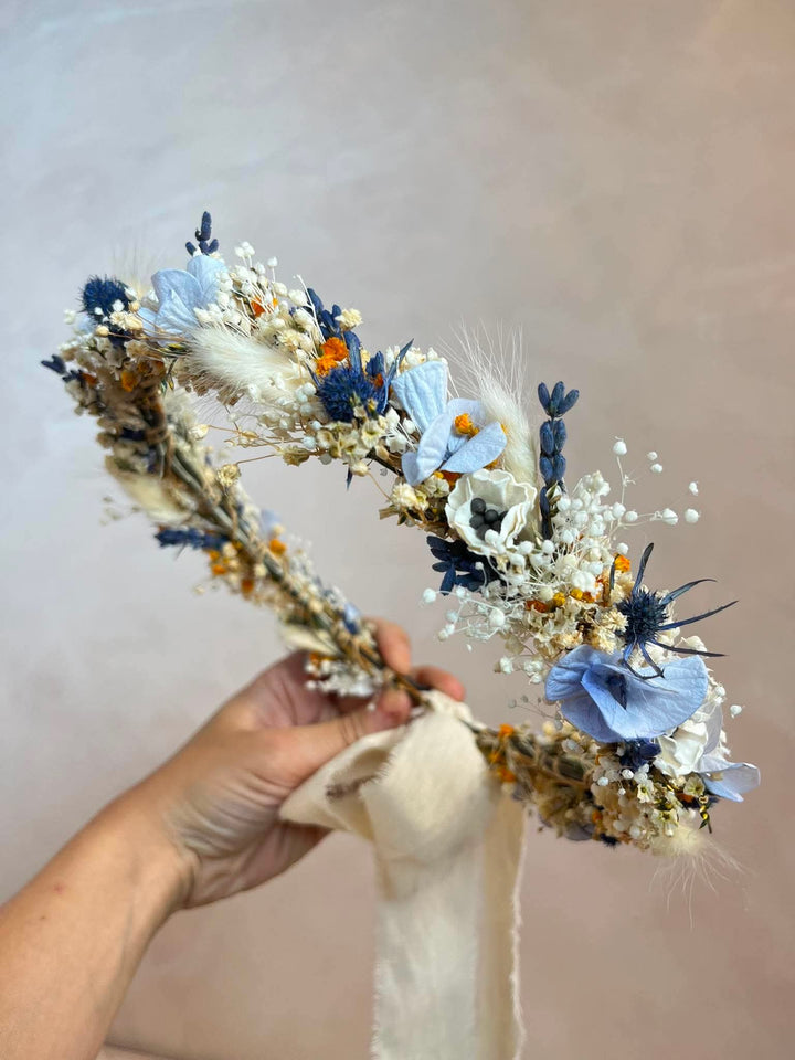 A dried floral headband with blue flowers, white baby's breath, and beige grass elements, held by a hand against a neutral beige background.