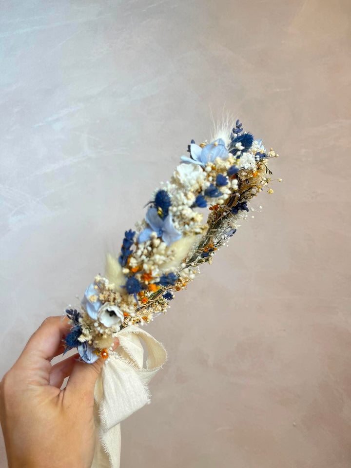 A hand holding a dried flower crown featuring light blue, navy, and white flowers with tiny gold accents and a cream ribbon, set against a soft beige background.