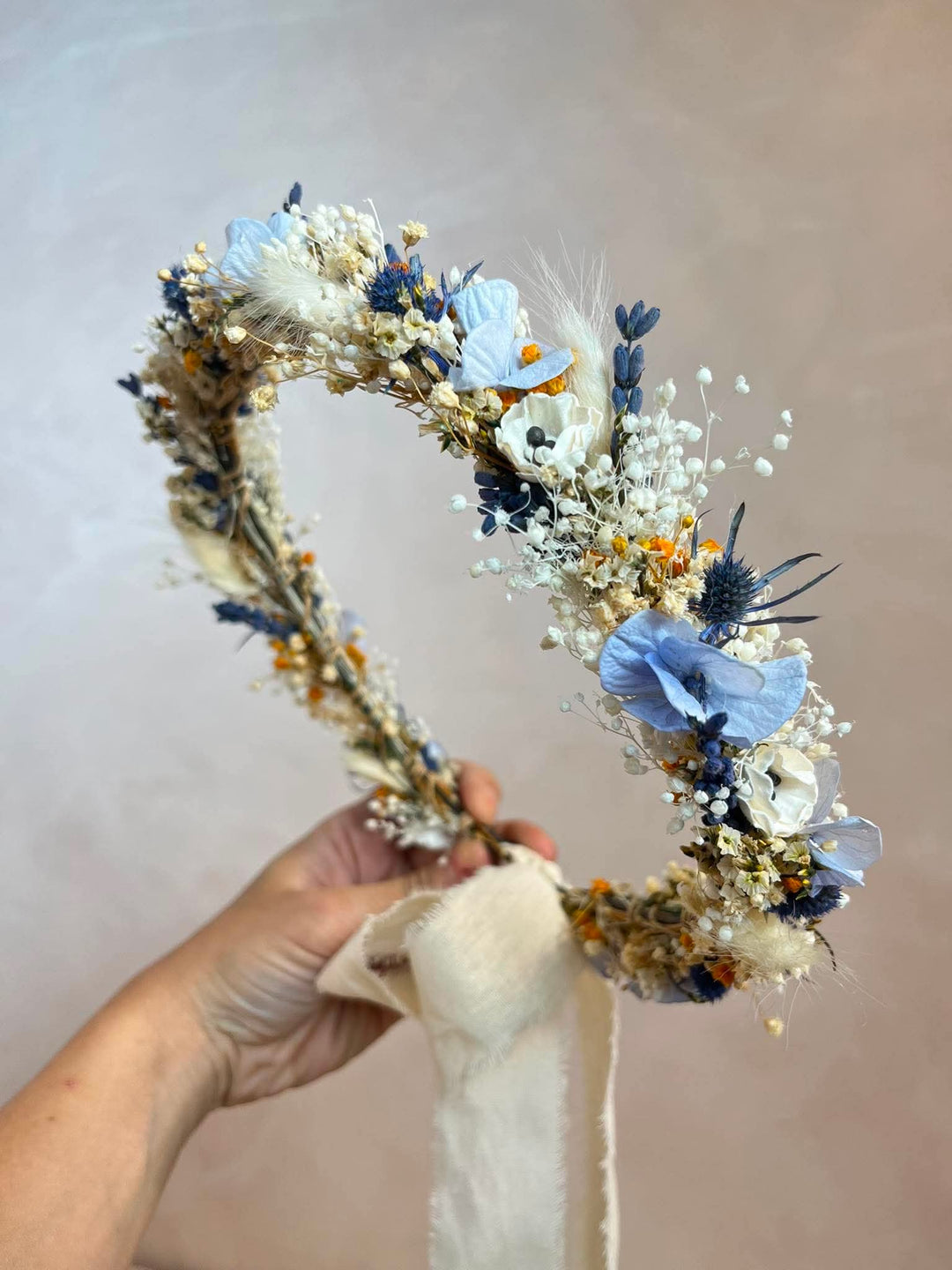 A dried flower crown held by a hand, featuring light blue, white, and beige botanical elements against a soft beige background.