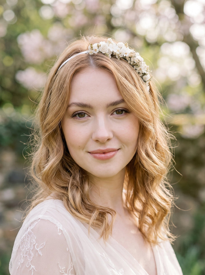 A woman with wavy blonde hair wearing a delicate white floral crown headband and a light-colored lace top, photographed outdoors against a soft, blurred background of green foliage and pink blossoms.