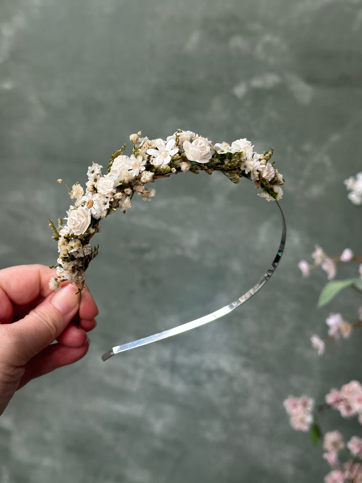 A hand holding a silver metal headband decorated with small white and ivory  flowers and green moss, set against a textured gray background with soft pink floral bokeh.