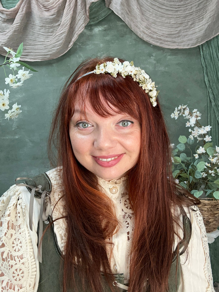 A woman with long auburn hair and a white flower crown lies on a dark green textured surface, wearing a cream lace top. White flowers and green leaves are scattered around her.