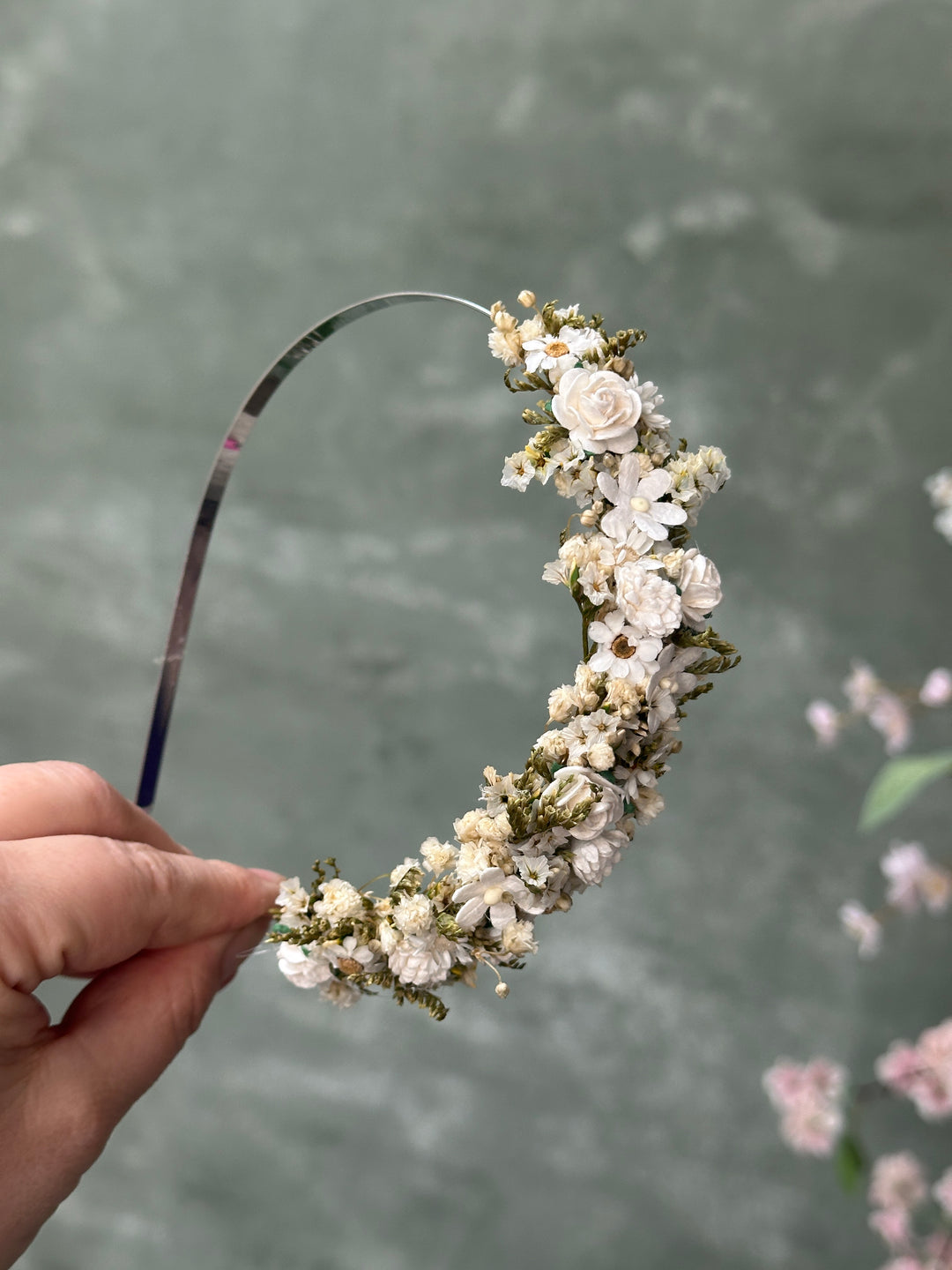 A hand holding a curved headband decorated with small white and cream flowers and green foliage, set against a soft gray textured background.