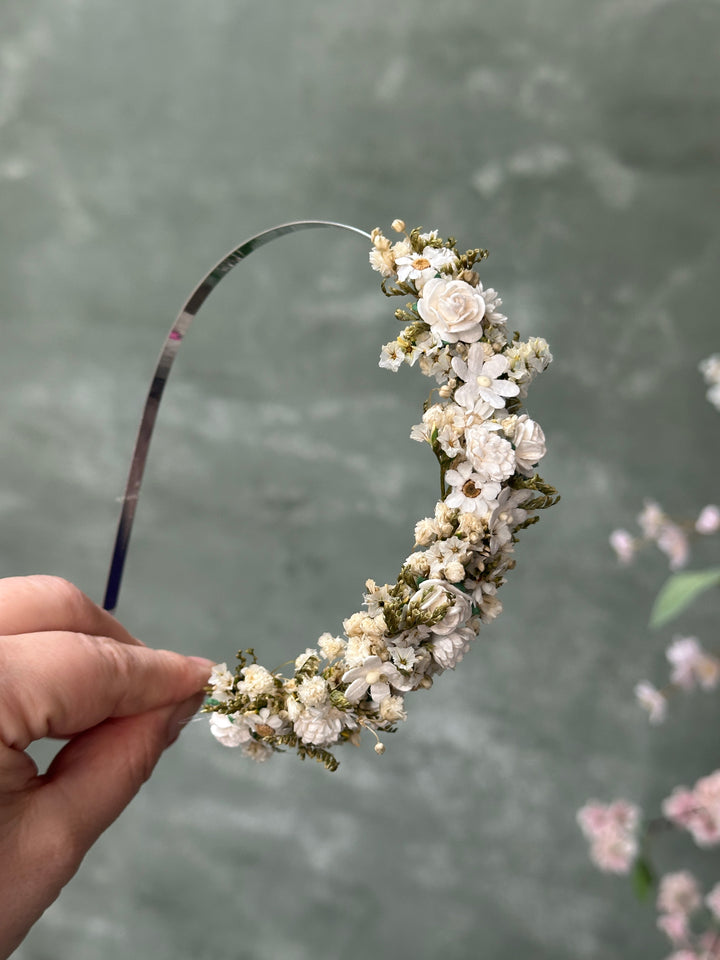 A hand holding a curved headband decorated with small white and cream flowers and green foliage, set against a soft gray textured background.