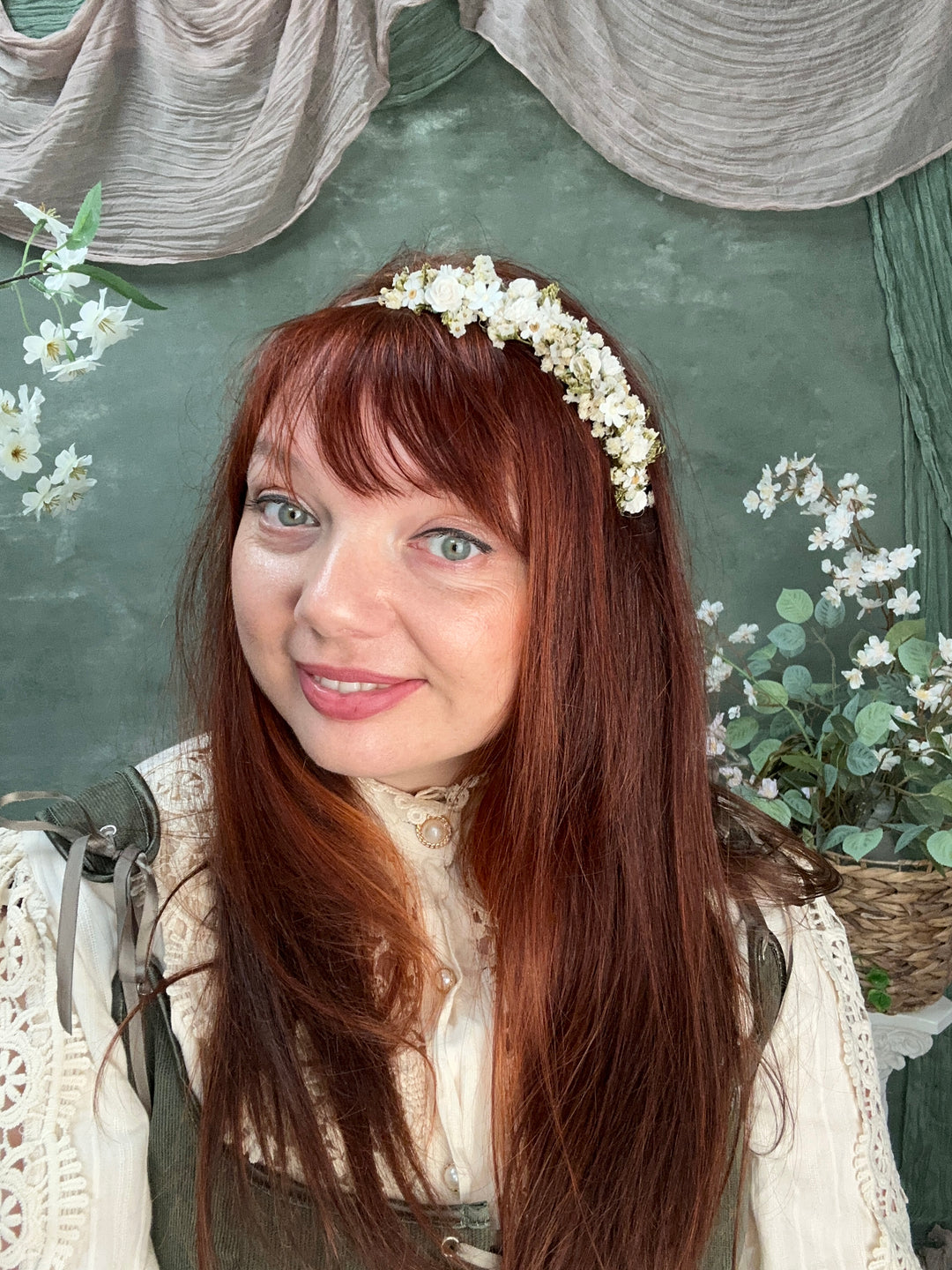 A woman with long auburn hair wearing a delicate white floral crown, photographed in a styled setting with a dark green textured backdrop, white flowers, and soft neutral fabric props.