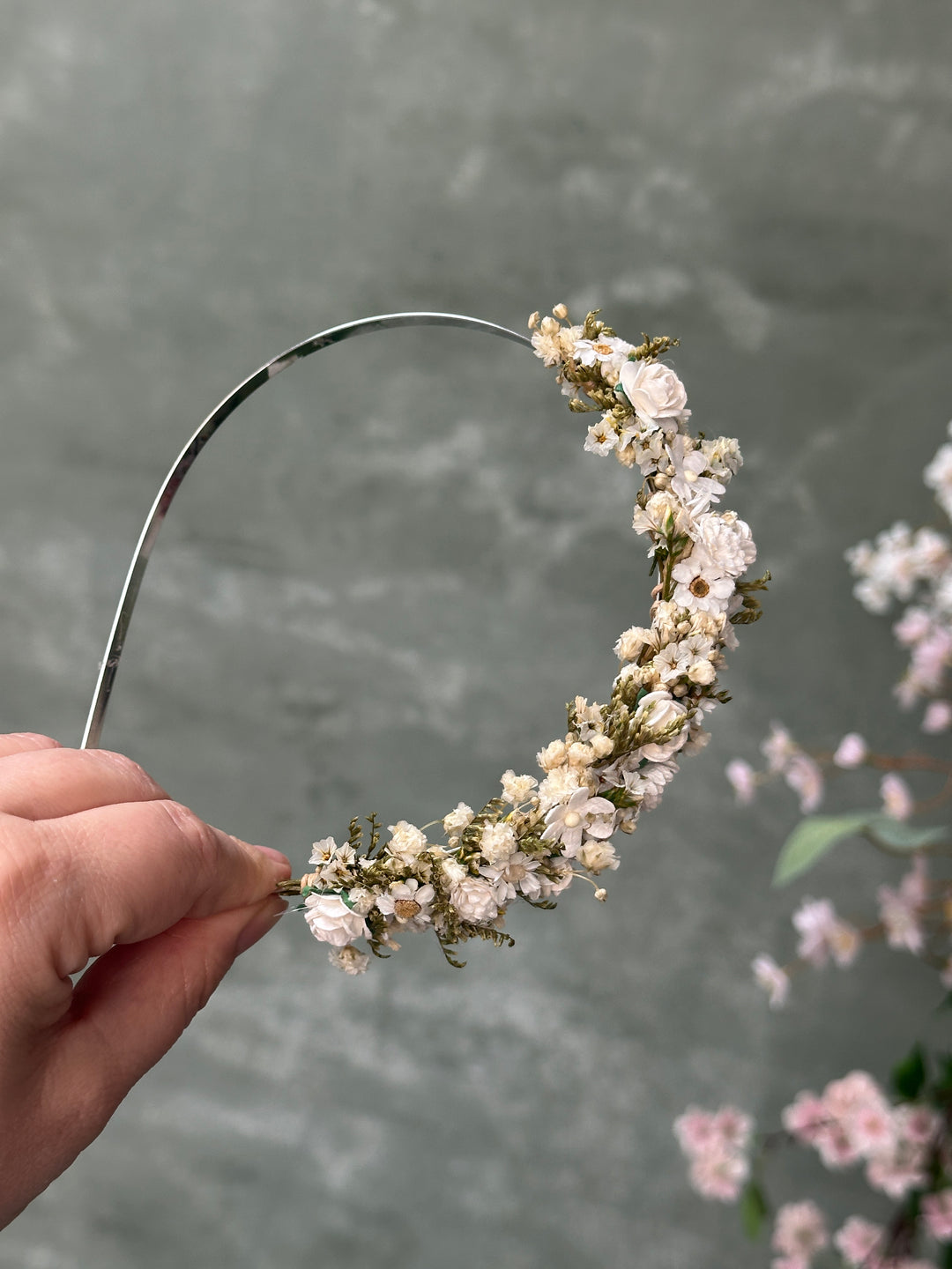 A hand holding a silver metal headband decorated with a dense wreath of small white and cream flowers and green foliage, set against a soft gray textured background with blurred pink flowers.