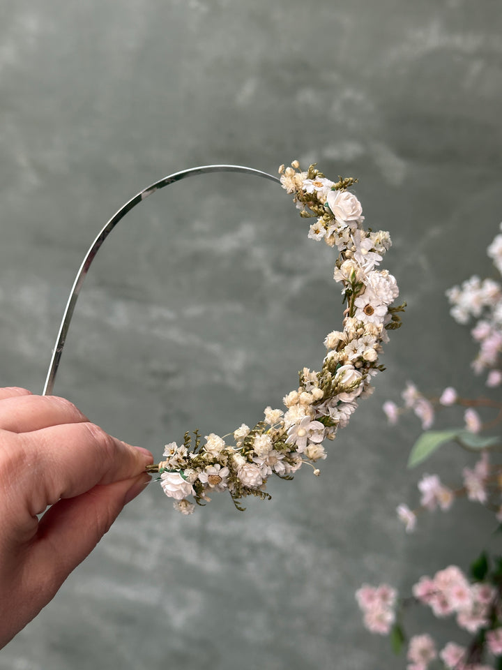 A hand holding a silver metal headband decorated with a dense wreath of small white and cream flowers and green foliage, set against a soft gray textured background with blurred pink flowers.