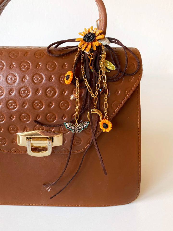 A brown textured leather handbag with a gold-tone clasp, decorated with a dark brown ribbon and autumn-themed charms including a sunflower, butterfly, and moon pendant.