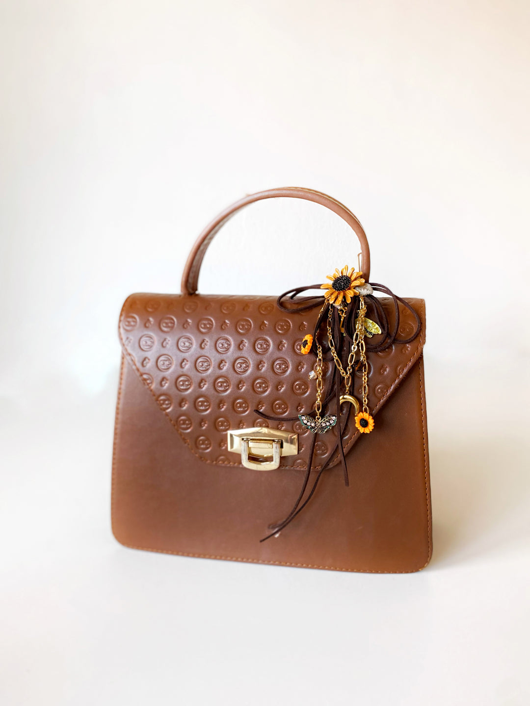 A brown leather handbag with an embossed circular pattern on the front flap, featuring a top handle, gold-tone hardware, and a decorative hanging charm with yellow and orange floral details.