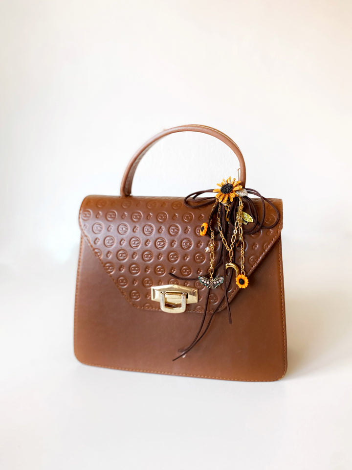 A brown leather handbag with an embossed circular pattern on the front flap, featuring a top handle, gold-tone hardware, and a decorative hanging charm with yellow and orange floral details.