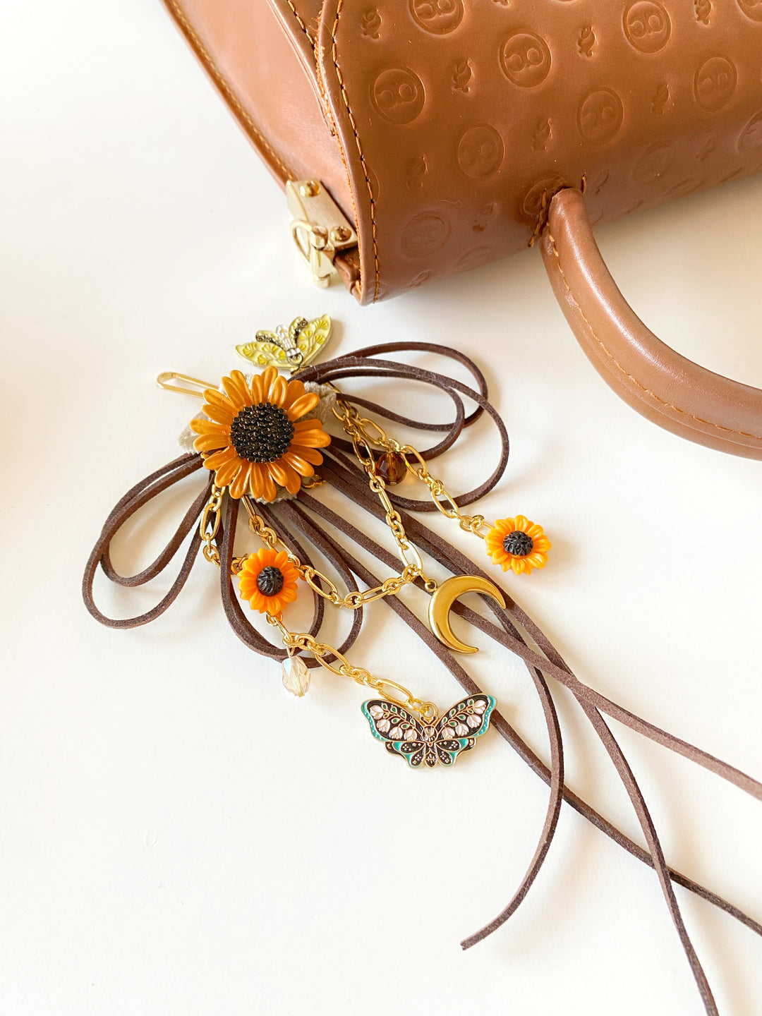 A close-up flat lay of a brown leather handbag handle and a decorative accessory with dangling chains, sunflower charms, a butterfly charm, and a crescent moon charm on a white background.