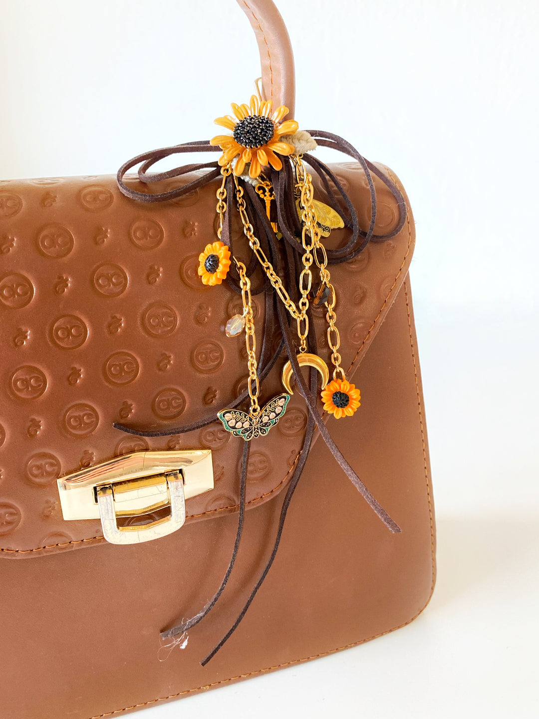 A close-up of a brown textured leather handbag with a gold clasp, decorated with a brown ribbon bow, gold chains, and sunflower charms.